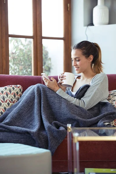 Ładna młoda kobieta używała swojego telefonu komórkowego podczas picia kawy na kanapie w domu. — Zdjęcie stockowe