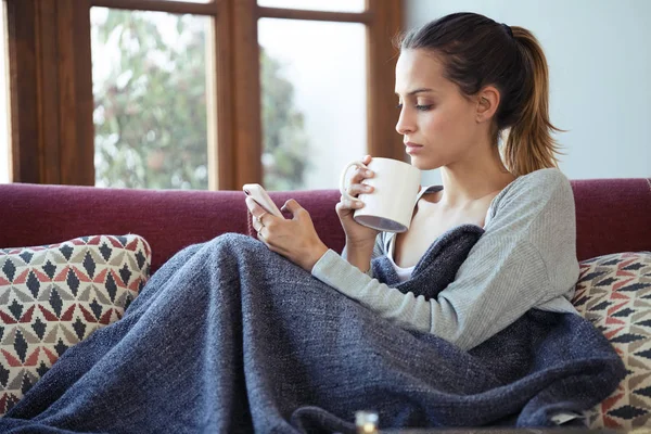 Hübsche junge Frau benutzt ihr Handy beim Kaffeetrinken zu Hause auf dem Sofa. — Stockfoto