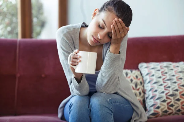 Depressive junge Frau mit Kopfschmerzen beim Kaffeetrinken auf dem heimischen Sofa. — Stockfoto