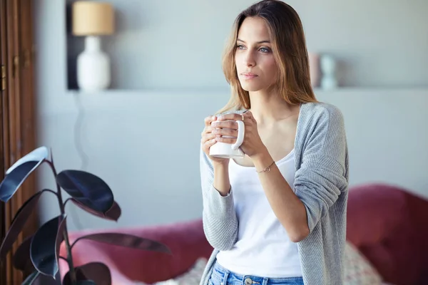 Schöne junge Frau, die am Fenster steht und zu Hause im Wohnzimmer Kaffee trinkt. — Stockfoto