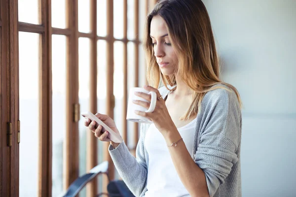 Hermosa mujer joven usando su teléfono móvil mientras bebe café cerca de la ventana en la sala de estar en casa . — Foto de Stock