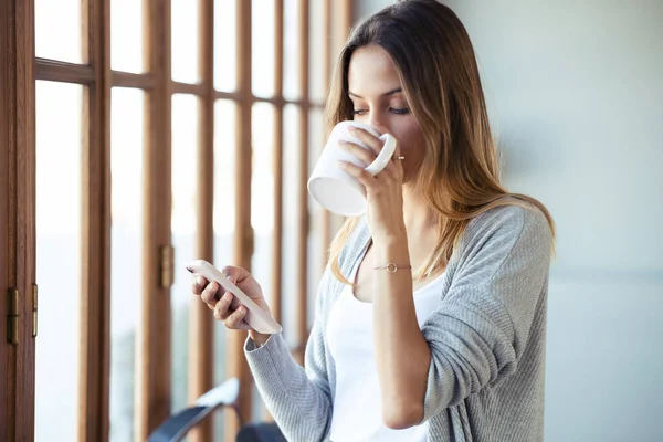 Hermosa mujer joven usando su teléfono móvil mientras bebe café cerca de la ventana en la sala de estar en casa . — Foto de Stock