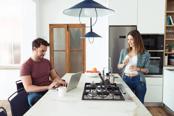 Schöner junger Mann, der seinen Laptop benutzt und seine Freundin Joghurt isst, während sie zu Hause in der Küche steht. — Stockfoto