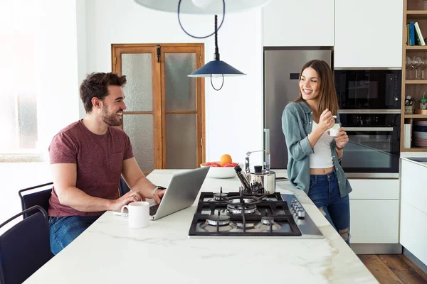 Bonito jovem usando seu laptop e sua namorada comendo iogurte enquanto estava na cozinha em casa . — Fotografia de Stock