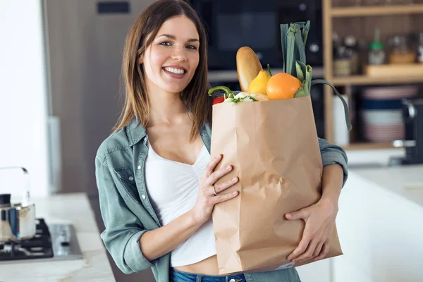 Gyönyörű fiatal nő nézi a kamerát, miközben bevásárlótáska friss zöldségek a konyhában otthon. — Stock Fotó