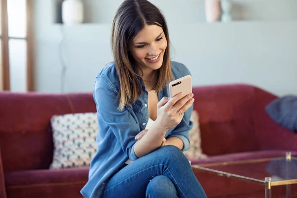 Mujer bastante joven usando su teléfono móvil mientras está sentada en el sofá en casa . — Foto de Stock