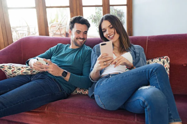 Preciosa pareja joven usando su teléfono móvil mientras se sienta en el sofá en casa . —  Fotos de Stock