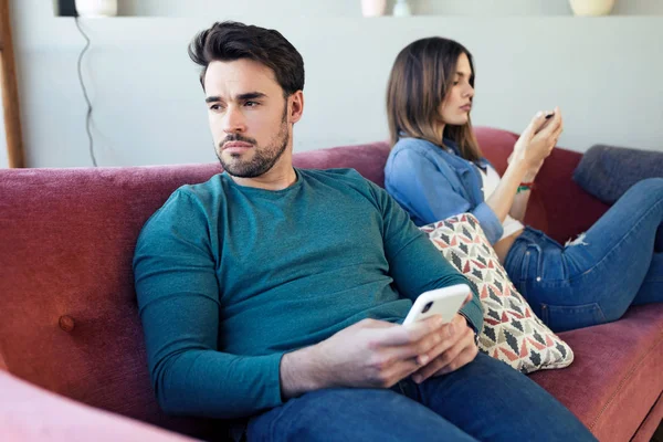 Irritado jovem casal ignorando uns aos outros usando o telefone depois de uma discussão enquanto sentado no sofá em casa . — Fotografia de Stock