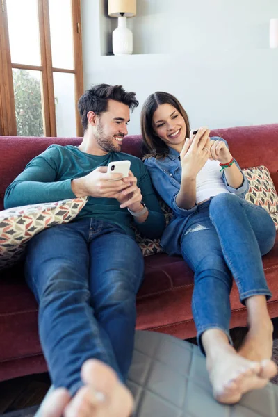 Schönes junges Paar benutzt sein Handy, während es zu Hause auf dem Sofa sitzt. — Stockfoto