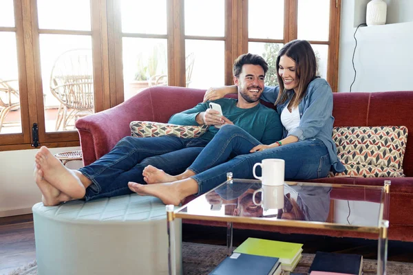 Schönes junges Paar benutzt sein Handy, während es zu Hause auf dem Sofa sitzt. — Stockfoto