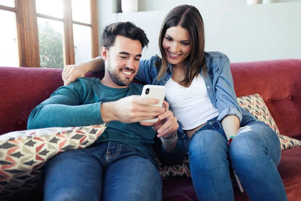 Прекрасная молодая пара с помощью мобильного телефона, сидя дома на диване . — стоковое фото