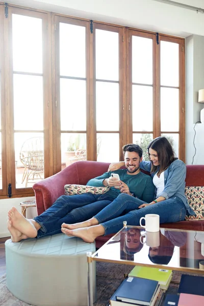 Schönes junges Paar benutzt sein Handy, während es zu Hause auf dem Sofa sitzt. — Stockfoto