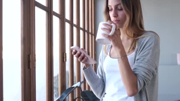 自宅のリビングルームの窓の近くでコーヒーを飲みながら携帯電話を使う美しい若い女性のビデオ — ストック動画