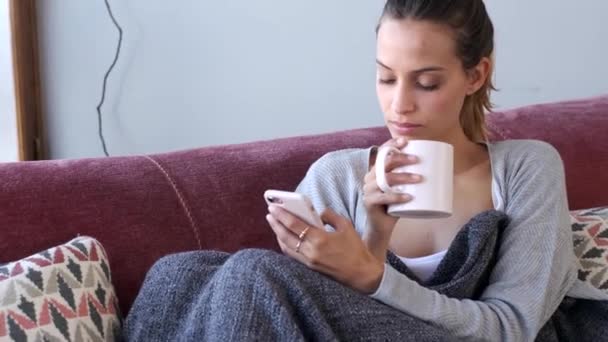 自宅のソファでコーヒーを飲みながら携帯電話を使用してかなり若い女性のビデオ — ストック動画
