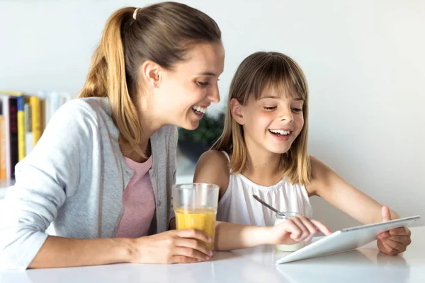 Bastante joven madre y su hija usando tableta digital mientras desayuna en casa . — Foto de Stock