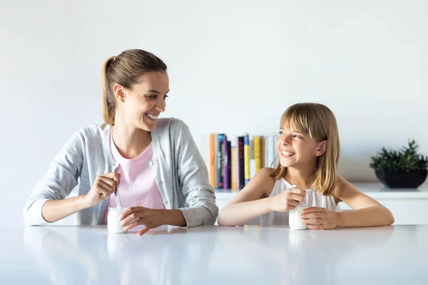 Ładna młoda matka i jej córka patrząc na siebie podczas jedzenia jogurtu w domu. — Zdjęcie stockowe