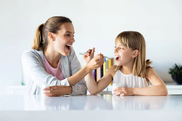 Bela mãe e filha alimentando iogurte uns aos outros em casa . — Fotografia de Stock