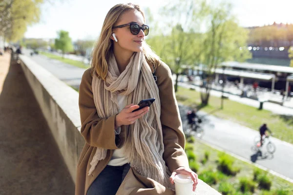 Mujer bastante joven escuchando música con auriculares inalámbricos y el teléfono inteligente en la calle . — Foto de Stock