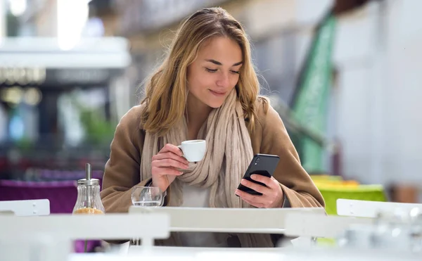 Mulher bonita usando seu telefone celular enquanto bebe uma xícara de café no terraço de um café . — Fotografia de Stock