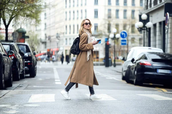Pretty ung kvinna korsar gatan medan du håller smartphone och tittar i sidled. — Stockfoto