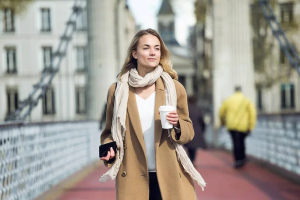 Krásná mladá žena, která při chůzi na ulici drží šálek kávy a smartphone. — Stock fotografie
