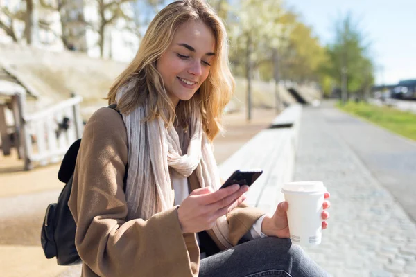 Mooie jonge vrouw met behulp van haar mobiele telefoon terwijl het houden van kopje koffie in de straat. — Stockfoto