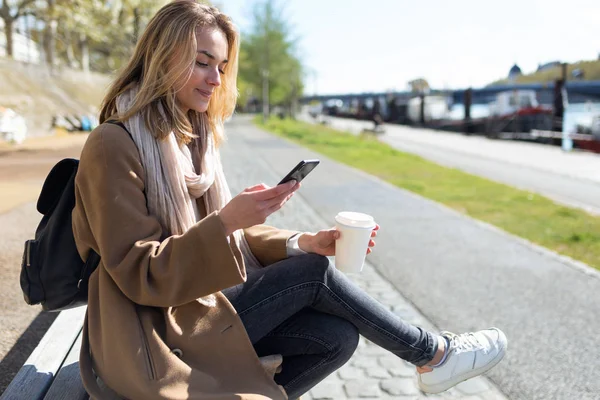 Bella giovane donna che usa il suo telefono cellulare mentre tiene in mano una tazza di caffè per strada . — Foto Stock