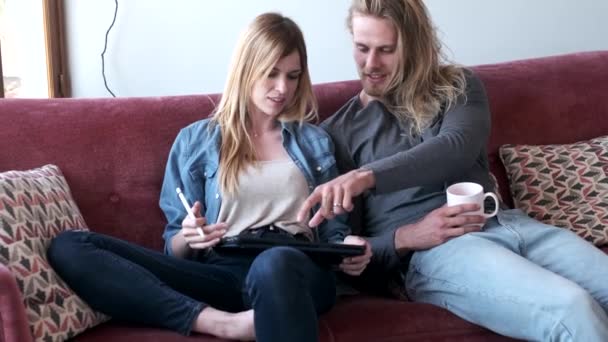 Beau jeune couple dessin avec ils tablette numérique tout en étant assis sur le canapé à la maison . — Video