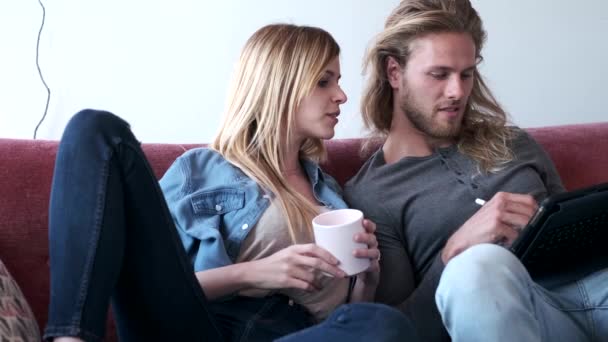 Beau jeune couple dessin avec ils tablette numérique tout en étant assis sur le canapé à la maison . — Video
