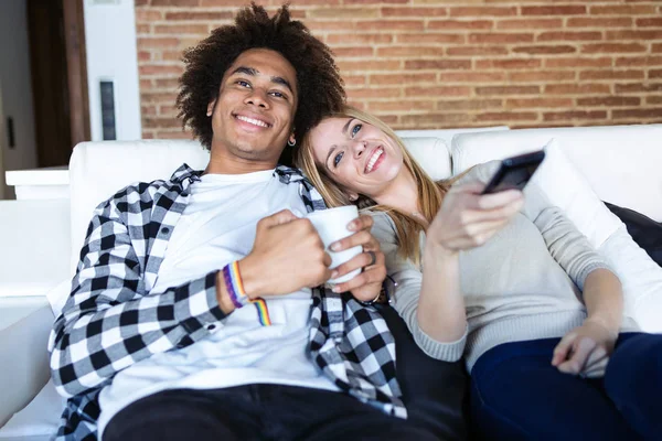 Relajada pareja joven cambiando de canal con el mando a distancia mientras ve la televisión en el sofá en casa . — Foto de Stock