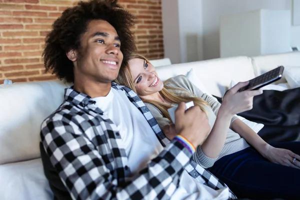 Relaxado jovem casal mudando de canais com o controle remoto enquanto assiste TV no sofá em casa . — Fotografia de Stock
