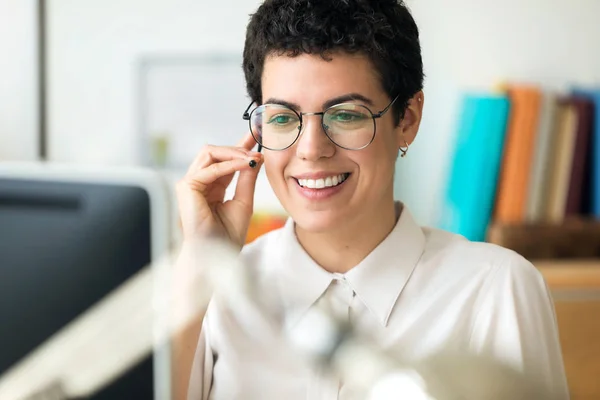 Sorrindo jovem mulher de negócios usando suas mãos livres para fazer uma chamada enquanto trabalhava com seu computador no escritório . — Fotografia de Stock