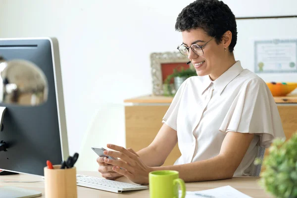 Усміхнена молода бізнес-леді використовує свій смартфон під час роботи з ноутбуком в офісі . — стокове фото