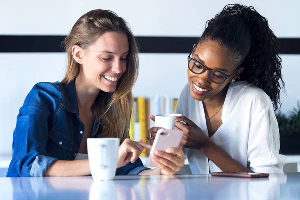 Two pretty young business women using her mobile phone together while taking a break in the office. — Stock Photo, Image
