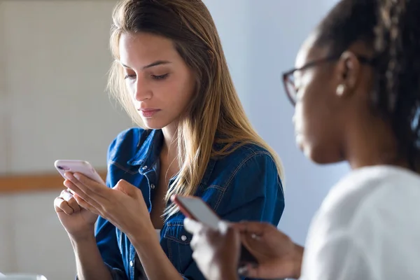 Dos mujeres de negocios bastante jóvenes usando sus teléfonos móviles en la oficina . — Foto de Stock