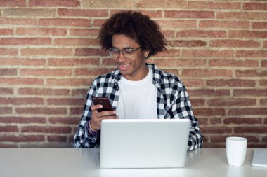 Evde dizüstü bilgisayar ile çalışırken onun akıllı telefon kullanarak gözlük ile Yakışıklı genç afro-amerikan adam.