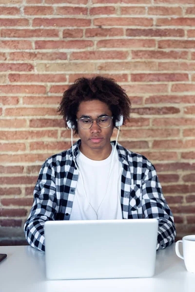 Knappe jonge Afro-Amerikaanse man met brillen luisteren naar muziek met een koptelefoon tijdens het gebruik van zijn laptop thuis. — Stockfoto