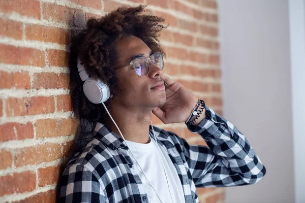 Jovem afro-americano relaxado ouvindo música com fones de ouvido apoiados na parede de pedra em casa . — Fotografia de Stock