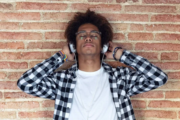 Ontspannen Afro-Amerikaanse jonge man luisteren naar muziek met hoofdtelefoon leunend op stenen muur thuis. — Stockfoto