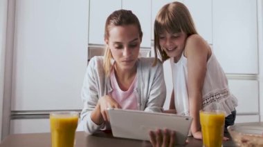 Evde mutfakta kahvaltı yaparken dijital tablet kullanarak Pretty genç anne ve kızı.