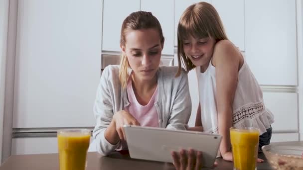 Красивая молодая мать и ее дочь, используя цифровой планшет во время завтрака на кухне дома . — стоковое видео