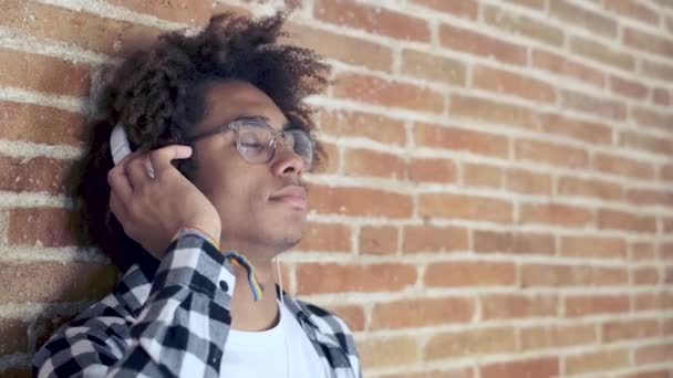 Joven afroamericano relajado escuchando música con auriculares apoyados en la pared de piedra en casa . — Vídeos de Stock