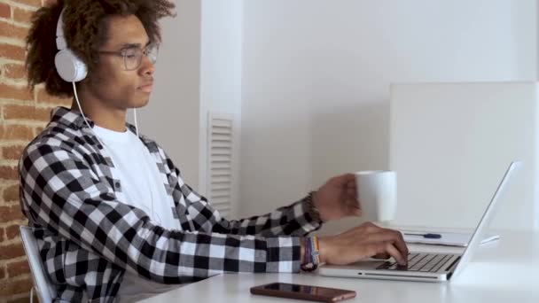 Beau jeune homme afro-américain boire du café tout en travaillant avec un ordinateur portable à la maison . — Video