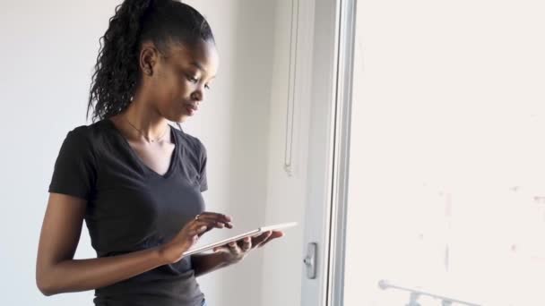 Jolie jeune femme utilisant sa tablette numérique et regardant la caméra à côté de la fenêtre à la maison . — Video