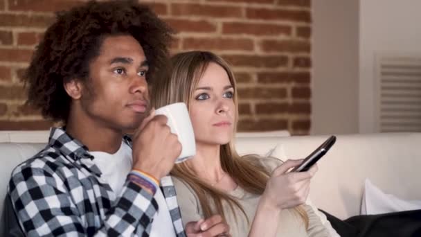 Jovem casal feliz mudando de canais com o controle remoto enquanto assiste TV no sofá em casa . — Vídeo de Stock