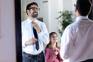 Handsome young father fixing his tie in front of the mirror and his pretty daughter copying it at home. clipart
