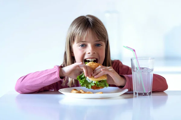 Pretty little girl eating a complete hamburger while looking at camera at home. — Stock Photo, Image