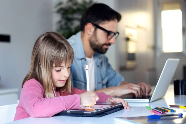 Linda niña dibujando en su tableta digital mientras su padre trabaja con el ordenador portátil en casa . — Foto de Stock