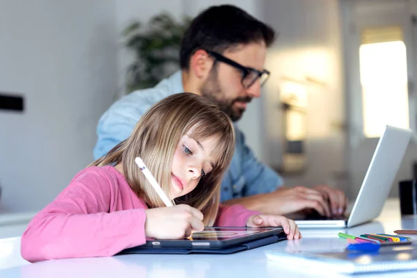 Linda niña dibujando en su tableta digital mientras su padre trabaja con el ordenador portátil en casa . — Foto de Stock