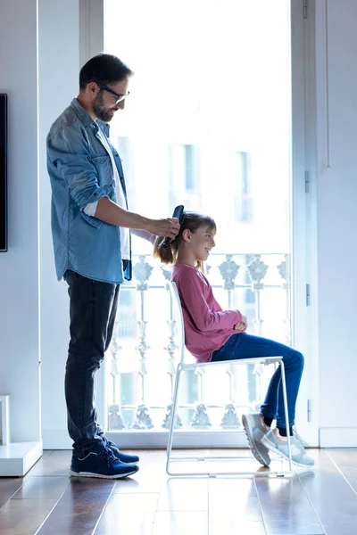 Bonito jovem pai fazendo um rabo de cavalo para sua filha enquanto ela se senta em uma cadeira em casa . — Fotografia de Stock